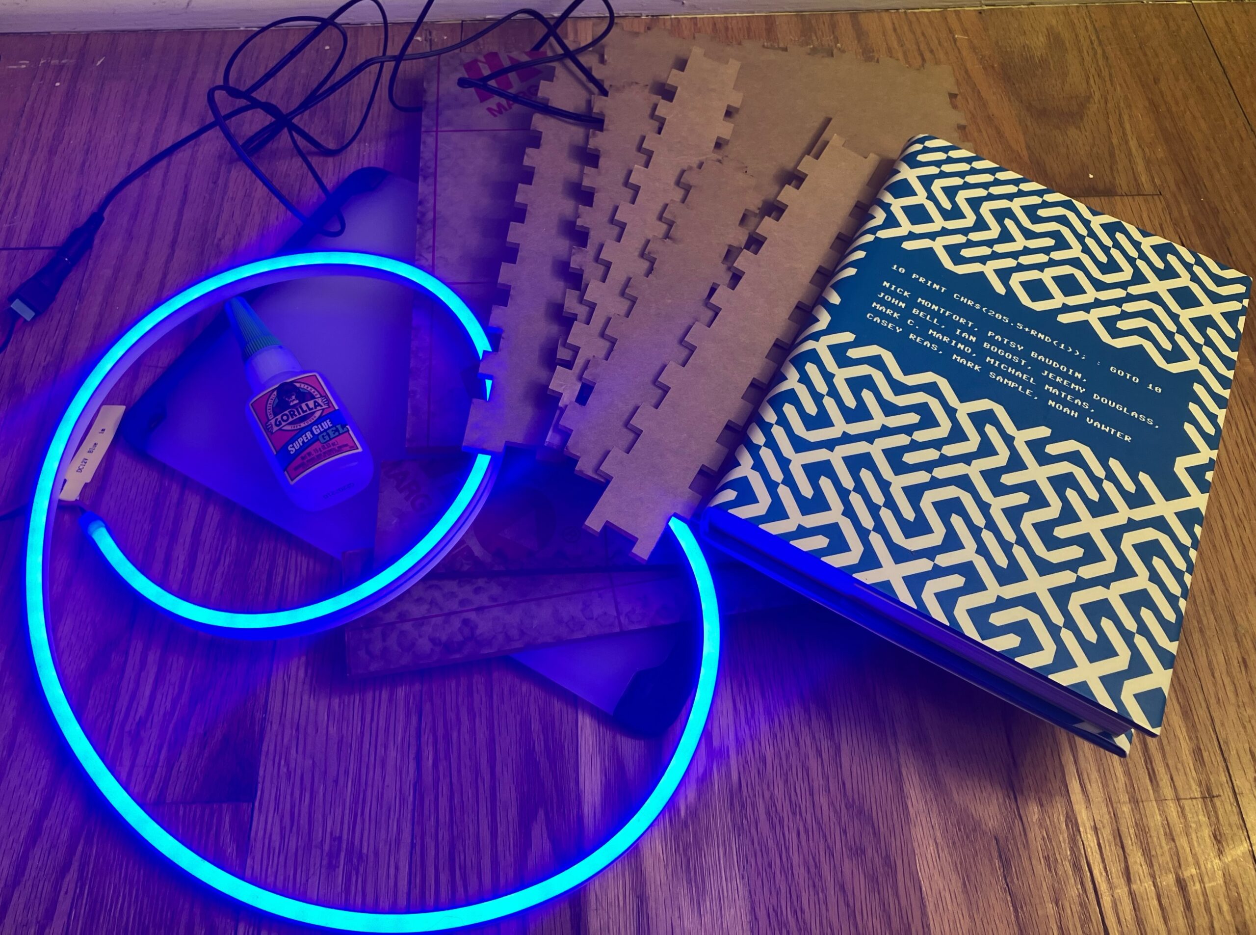 Photo of materials for the Ghost Books project artfully arranged on a floor, including a swirl of blue LEDs with silicone diffusion making them look like neon lights, superglue, acrylic and glass cut to size to be assembled into a rectangular-prism/book shape with smoothe or crenellated edges, and one of the books I'm basing the initial prototype on (10 PRINT) because of it's interesting blue and white patterned cover.