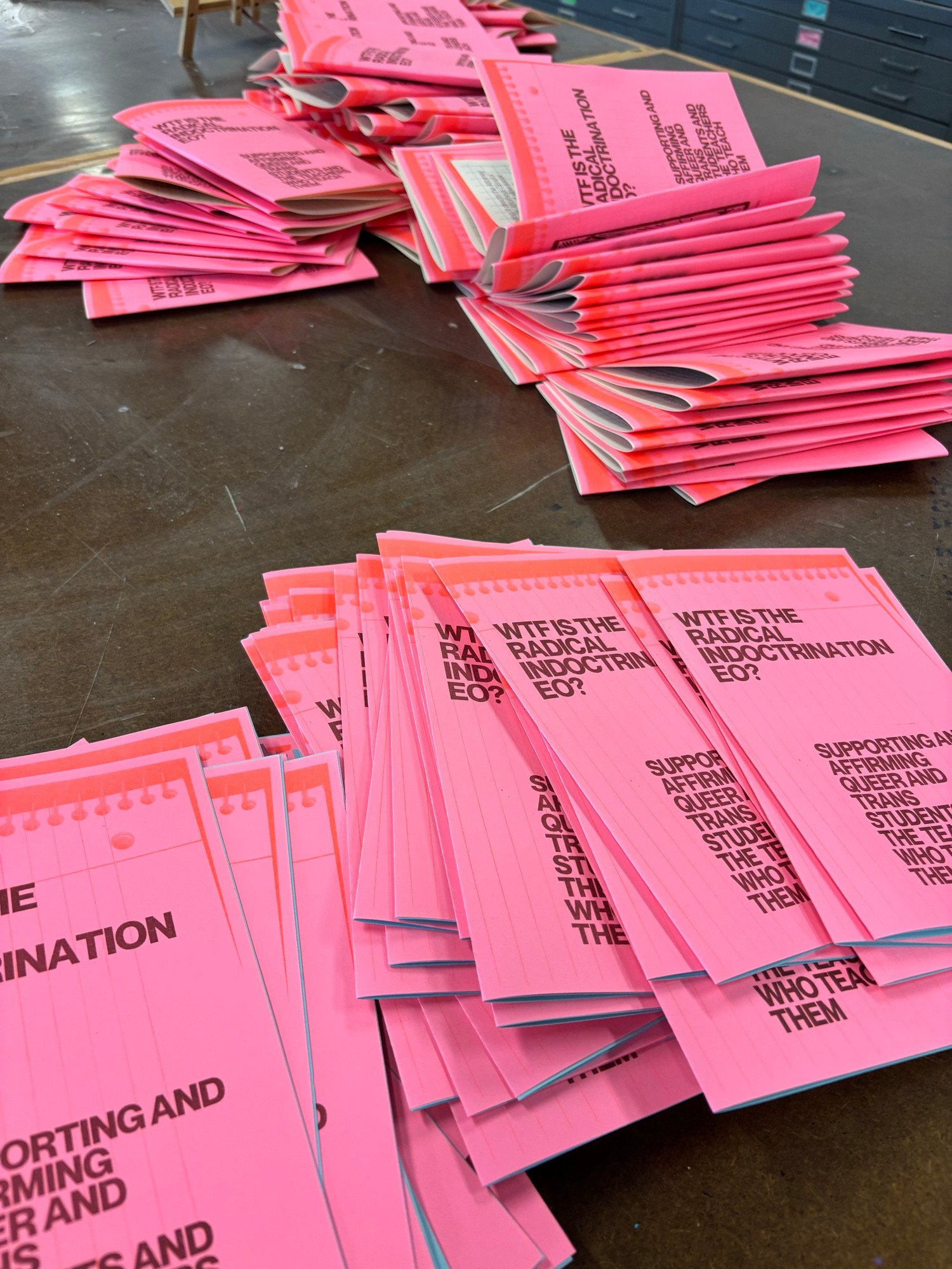 Photo of stacks of fluorescent zines with the text “WTF is the radical indoctrination EO? Supporting and affirming queer and trans students and the teachers who team them”